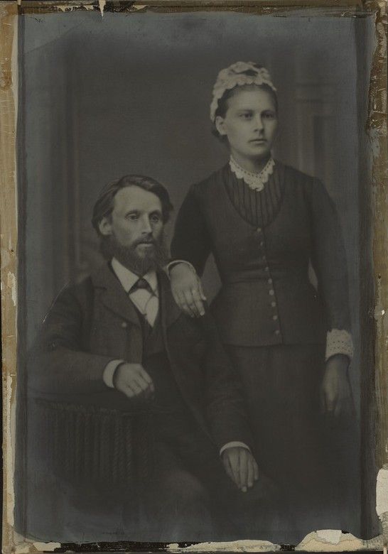Full-length studio portrait: a bearded man seated, a young woman in a buttoned bodice and lace cap standing beside him with her hand on his shoulder.