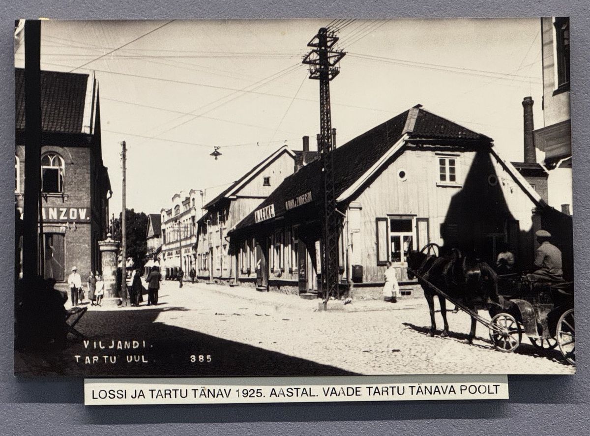Street view along Tartu street in 1925, with a horse and cart; a long low wooden building with the sign “H. LEOKE.” beyond a corner house with a round window.